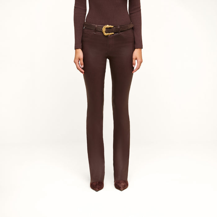 A person wearing a fitted brown long-sleeve top, the Ruth Coated Straight-Leg Jean with a gold-buckled belt, and pointed brown shoes stands against a plain white background.