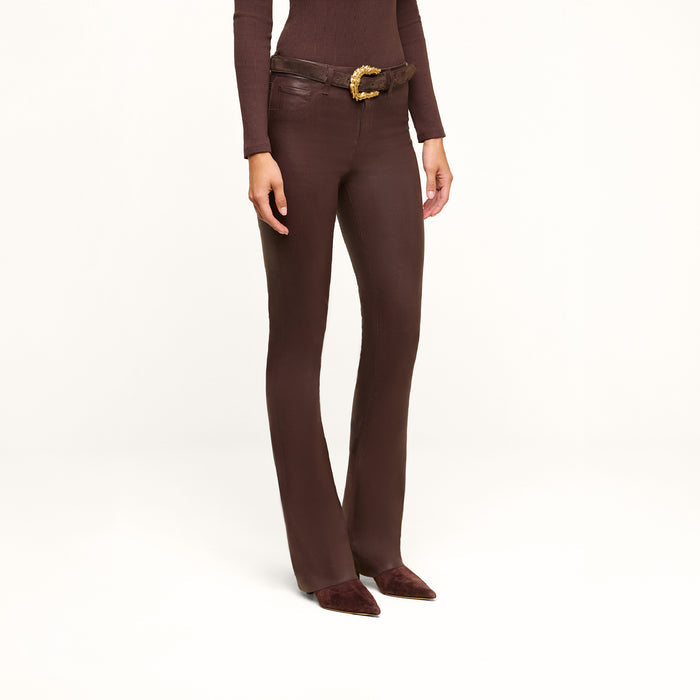 Wearing the Ruth Coated Straight-Leg Jean in brown, along with a coordinating brown long-sleeve top, a brown belt featuring a gold buckle, and pointed brown shoes, a person stands against a plain white background.