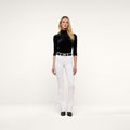 A woman stands facing forward, wearing a black turtleneck top, the Ruth Straight-Leg Jean in white with a black belt, and black shoes, posed against a plain white background.