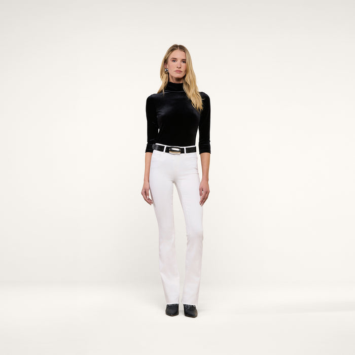 A woman stands facing forward, wearing a black turtleneck top, the Ruth Straight-Leg Jean in white with a black belt, and black shoes, posed against a plain white background.
