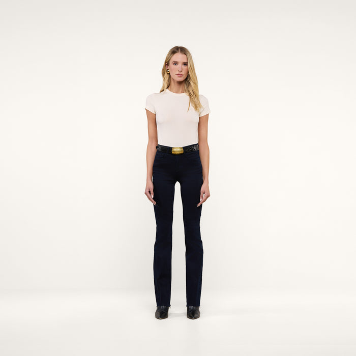 A woman stands facing forward against a plain white background, wearing a fitted white t-shirt, the Ruth Straight-Leg Jean in ultra dark indigo wash with a wide belt, and black shoes.