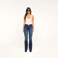A woman wearing a white tank top, Ruth Straight-Leg Jean in high-rise indigo wash, a black belt, black shoes, and sunglasses stands facing forward against a plain white background.