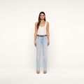 A woman stands facing forward against a plain white background, wearing a white tank top, the Ruth Straight-Leg Jean in light blue classic stretch denim with a raw hem, a belt, and beige heels.
