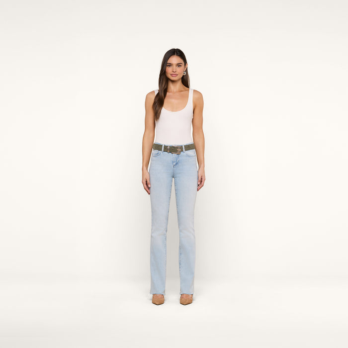A woman stands facing forward against a plain white background, wearing a white tank top, the Ruth Straight-Leg Jean in light blue classic stretch denim with a raw hem, a belt, and beige heels.