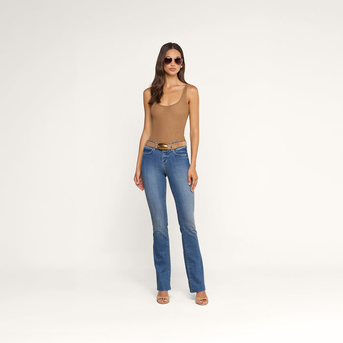 A woman wearing sunglasses, a tan sleeveless top, the Ruth Straight-Leg Jean in indigo with a raw hem, a brown belt, and heeled sandals stands against a plain white background.