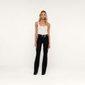 A woman with long blonde hair stands facing forward against a plain white background, wearing a light tank top, a black belt, black shoes, and high-rise black Ruth Straight-Leg Jeans.
