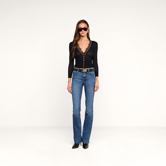 A woman wearing sunglasses, a black long-sleeve lace v-neck top, a belt, and black shoes stands against a plain white background. She completes her look with the Ruth Straight-Leg Jean in a medium indigo wash, crafted from stretch denim for a flattering fit.
