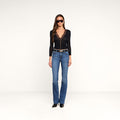 A woman wearing sunglasses, a black long-sleeve lace v-neck top, a belt, and black shoes stands against a plain white background. She completes her look with the Ruth Straight-Leg Jean in a medium indigo wash, crafted from stretch denim for a flattering fit.