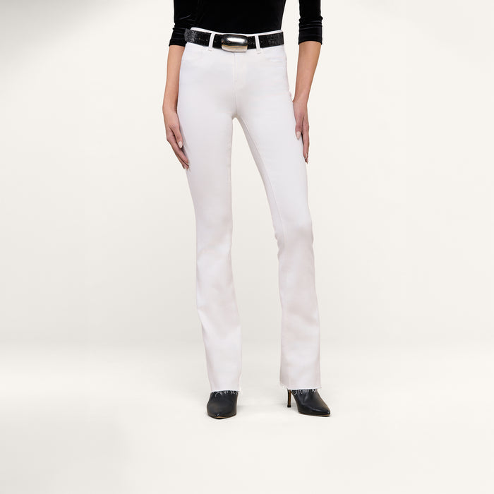 A person wearing the Ruth Straight-Leg Jean in white stretch denim, paired with a black top, a black belt with a silver buckle, and black pointed-toe heels stands against a plain white background.