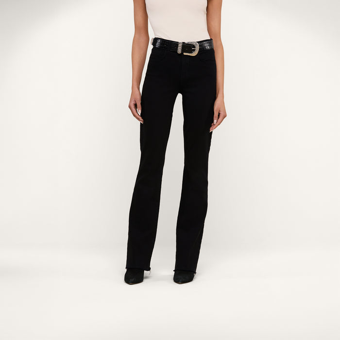 A person wearing the Ruth Straight-Leg Jean in black stretch denim, styled with a white sleeveless top, black shoes, and a black belt with a large buckle stands against a plain white background.
