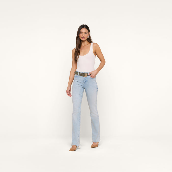 A woman stands against a plain white background wearing a white tank top, beige heels, and the Ruth Straight-Leg Jean in light blue classic stretch denim with a raw hem. She has long brown hair and a neutral expression, with one hand in her pocket.