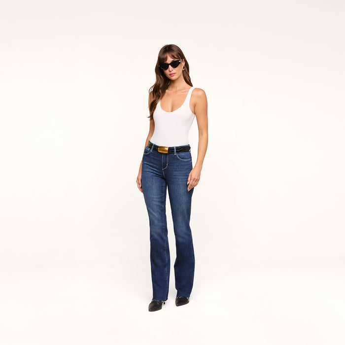 A woman wearing sunglasses, a white tank top, the Ruth Straight-Leg Jean in indigo wash, and black shoes stands against a plain white background, posing with one hand in her pocket and a relaxed posture.