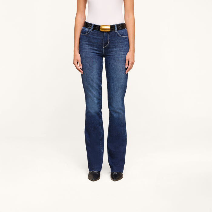 A person wearing a white sleeveless top, the Ruth Straight-Leg Jean in high-rise indigo wash, a black belt with a gold buckle, and black pointed shoes stands against a plain white background.
