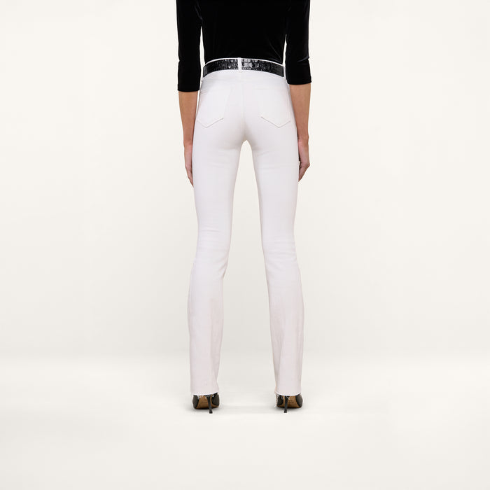 A person stands facing away against a plain white background, wearing the Ruth Straight-Leg Jean in white with a high-rise fit and slim straight leg, styled with a black belt, black long-sleeve top, and black-heeled shoes.