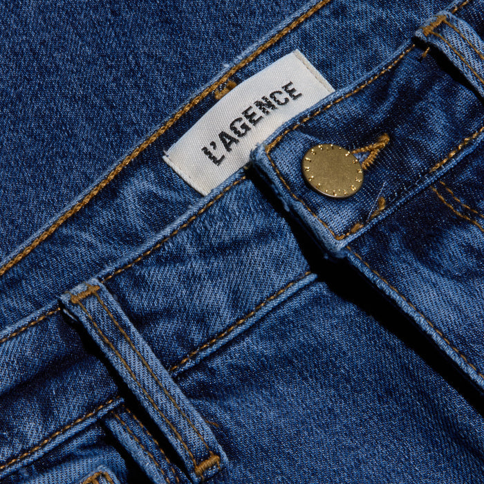 Close-up of blue stretch denim Ruth Straight-Leg Jean featuring a white LAGENCE label, a front belt loop, and a brass button. This fitted style comes in a medium indigo wash.