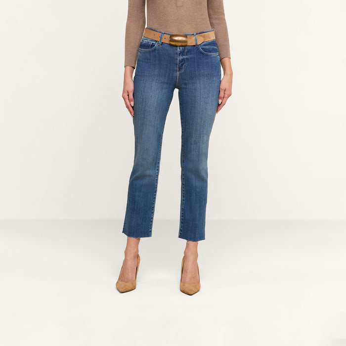 A person wearing a brown long-sleeve top, tan belt, and tan high heels stands against a plain white background, styled in blue Sada Cropped Slim-Leg Jeans with a raw hem.