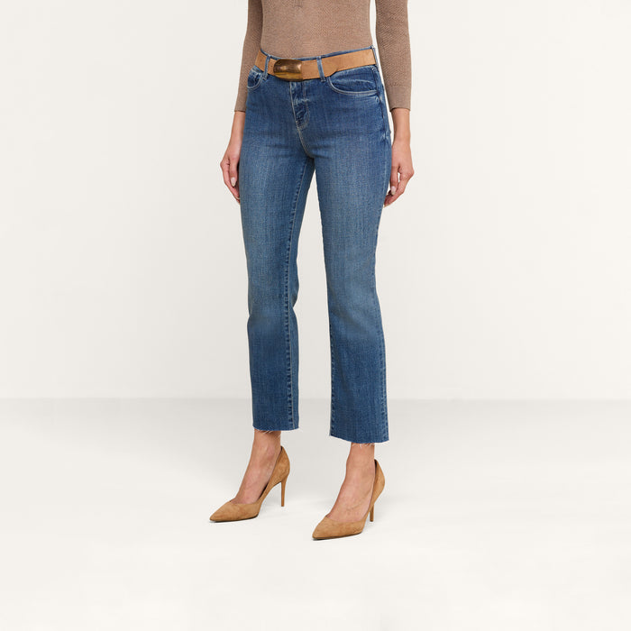 A person wearing a tan long-sleeve top, the Sada Cropped Slim-Leg Jean in blue with a raw hem and a brown belt, and tan high heels stands against a plain white background.