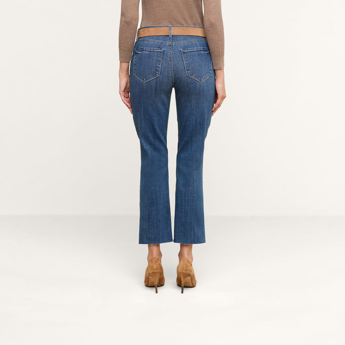 A person is shown from the back standing against a plain light background, wearing the Sada Cropped Slim-Leg Jean in denim blue with a raw hem, paired with a brown long-sleeve top and brown heeled shoes.
