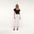 A woman stands against a plain white background, wearing sunglasses and posing with her hands by her sides. She is dressed in a black V-neck shirt, the Sada Slim-Leg Cropped Jean with stretch denim, a black belt, and black heels.