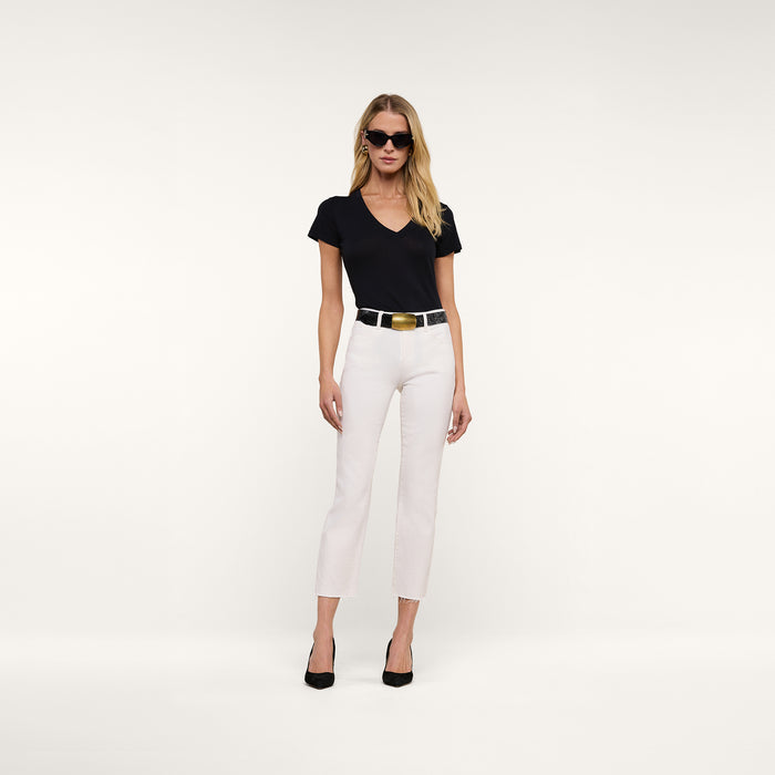 A woman stands against a plain white background, wearing sunglasses and posing with her hands by her sides. She is dressed in a black V-neck shirt, the Sada Slim-Leg Cropped Jean with stretch denim, a black belt, and black heels.