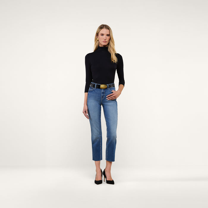 A woman stands against a plain background wearing a black turtleneck, black heels, and the Sada Slim-Leg Cropped Jean—a pair of blue high-rise cropped jeans crafted from premium stretch denim with a black belt. She has long blonde hair and her left hand rests in her pocket.