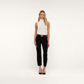 A woman stands against a plain white background, wearing a sleeveless white turtleneck top, the Sada Slim-Leg Cropped Jean in black with a belt, and black heels.