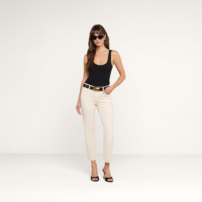 A woman wearing sunglasses, a black tank top, the Sada Slim-Leg Cropped Jean in high-rise white stretch denim, a black belt, and black heels stands against a plain white background.