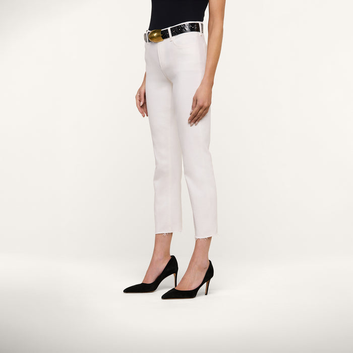 A woman stands against a white background wearing the Sada Slim-Leg Cropped Jean in stretch denim, paired with a black belt with a gold buckle, a black sleeveless top, and black high heels.