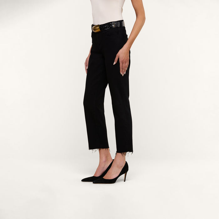 A person wearing a sleeveless white top, black Sada Slim-Leg Cropped Jeans with a frayed hem, a black belt, and black pointed-toe heels stands against a plain white background.