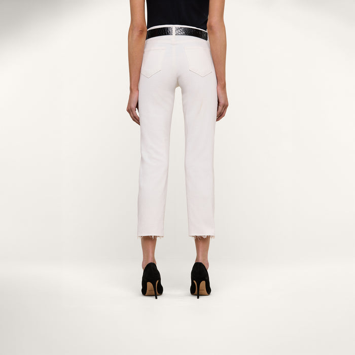 A person stands facing away, wearing the Sada Slim-Leg Cropped Jean—a high-rise white jean with frayed hems—paired with a black belt, black heels, and a dark top, against a plain white background.