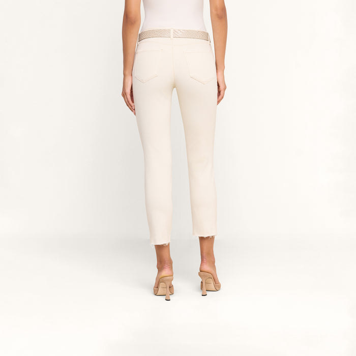 A woman stands facing away against a plain white background, wearing the Sada Slim-Leg Cropped Jean—off-white high-rise jeans crafted from premium stretch denim—paired with a white top, a beige woven belt, and beige heeled sandals.