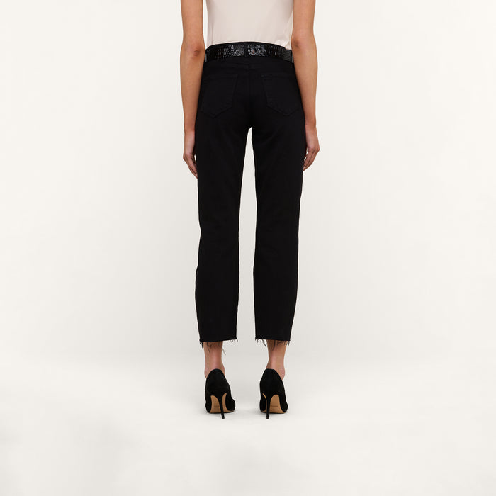 A person stands facing away, wearing the Sada Slim-Leg Cropped Jean—high-rise black jeans with frayed hems—paired with a white sleeveless top and black high-heeled shoes against a plain light background.