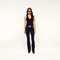A woman stands against a white background wearing a navy blue sleeveless top and pants, black shoes, a decorative belt, and dark sunglasses. Her Saffia Corduroy Pant in a high-rise slim-straight fit adds a contemporary touch to the outfit.