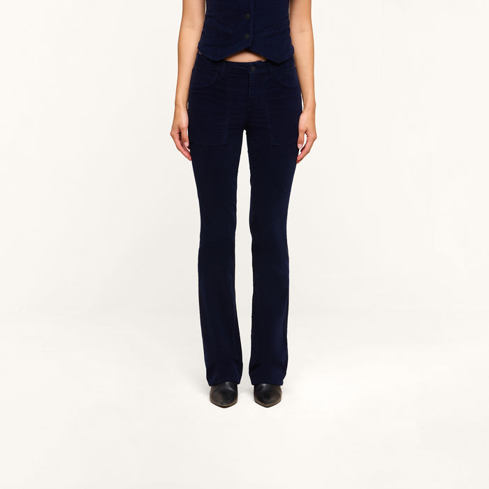 A person stands facing forward with their hands by their sides, wearing the Saffia Corduroy Pant in dark blue—a high-rise, flared style crafted from stretch corduroy—paired with a matching sleeveless vest against a plain white background. Their head is not visible in the image.