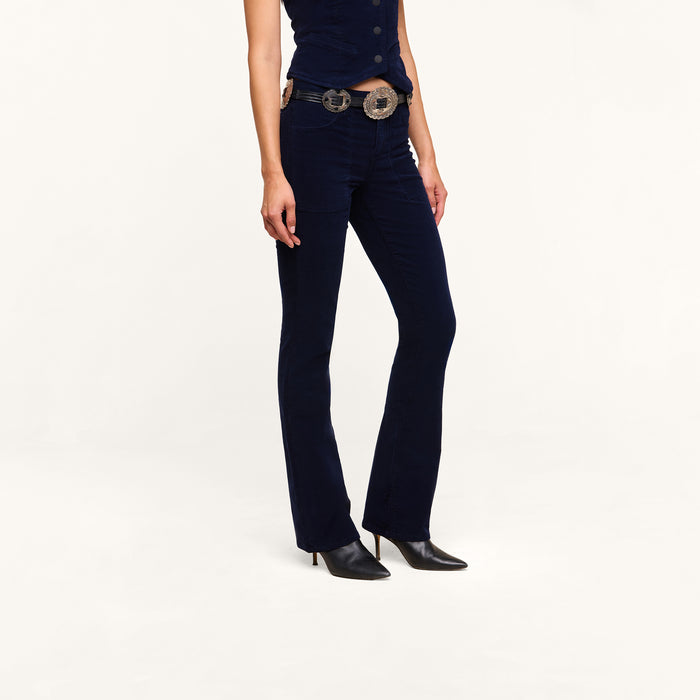 A person wearing fitted dark blue Saffia Corduroy Pants, a matching sleeveless top, a decorative belt with large buckles, and black pointed-toe shoes, standing against a plain white background.