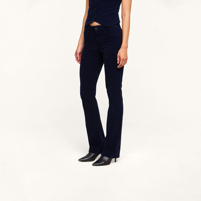A person wearing the Saffia Corduroy Pant in dark blue—styled as high rise, flared, and crafted from stretch corduroy—paired with a matching sleeveless top and black heeled boots stands against a plain white background. The person’s head is not visible.