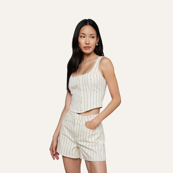 A woman poses against a plain light background, wearing the sleeveless cream Saga Top with nautical stripes and matching high-waisted shorts, one hand casually in her pocket.