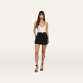 A woman stands against a plain white background wearing a white sleeveless top, the Saige Short in black stretch suiting, and black heeled sandals.