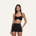 A woman wearing the Sally Bandeau Top paired with a matching high-waisted mini skirt with gold buttons stands against a plain, light background, channeling retro glamour.