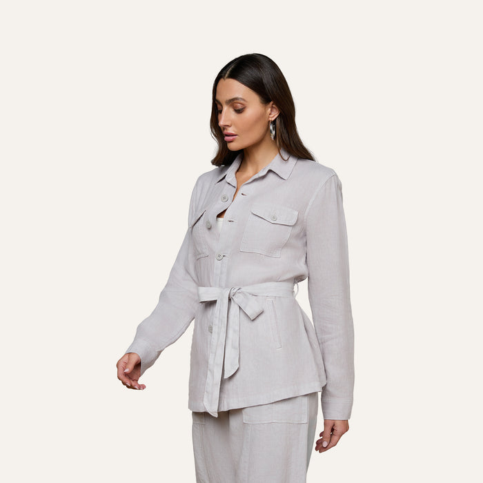 Wearing the Samaya Linen Jacket—a light-colored, versatile linen design with patch pockets—she pairs it with matching pants. Standing against a plain, off-white background, she looks down, her long dark hair cascading over her shoulders.