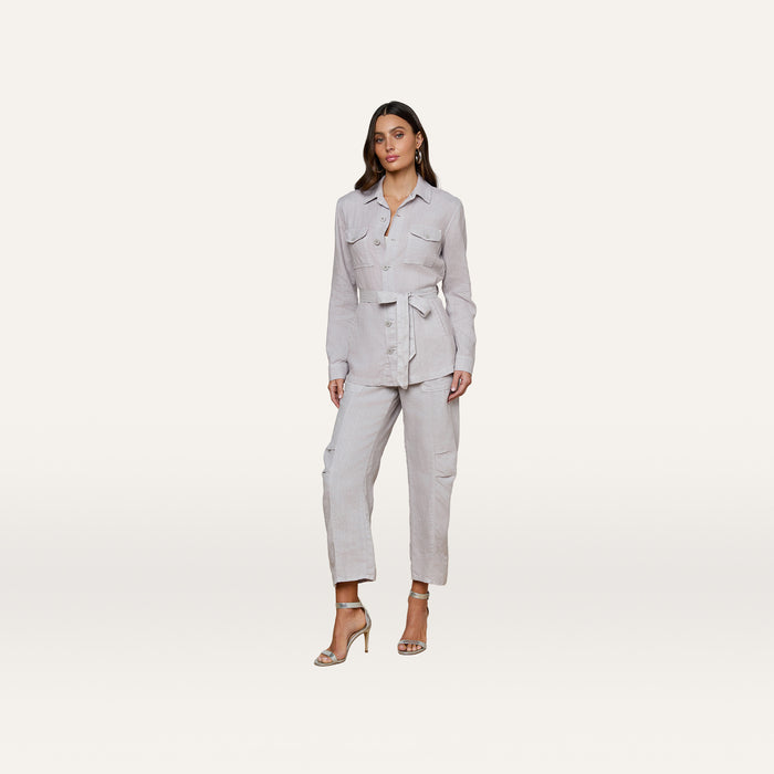 A woman stands facing forward against a plain white background, wearing the Samaya Linen Jacket in light gray, styled as a belted utility jumpsuit and paired with beige high-heeled sandals.