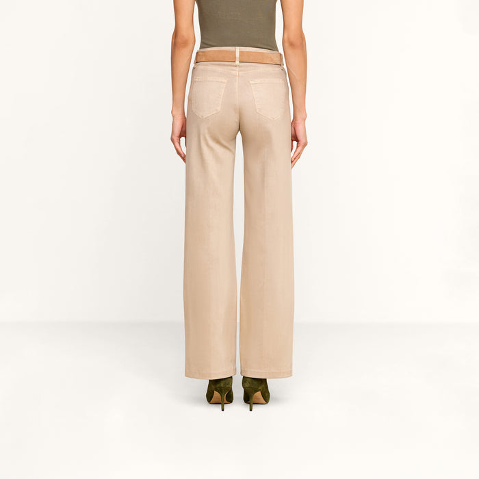 A person stands facing away, wearing the Scottie Coated Wide-Leg Jean in beige, paired with a tan belt, an olive green sleeveless top, and green heeled shoes against a plain white background.