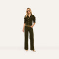 A woman wearing sunglasses, a dark green corduroy jacket, the Scottie Corduroy Pant in a matching high-rise wide-leg silhouette, a belt, and beige heels stands against a plain light background, capturing the essence of fall 2026 fashion.