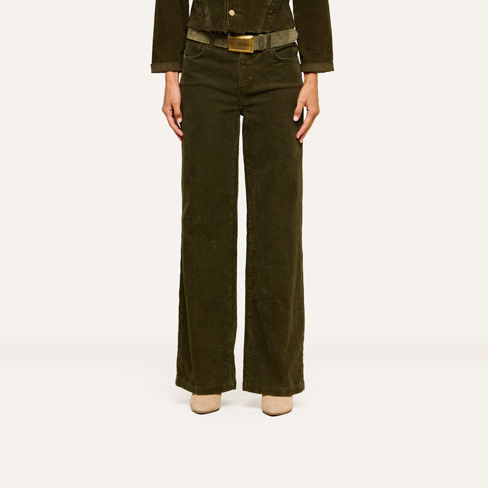 A person is standing against a plain white background, wearing the Scottie Corduroy Pant in dark green, paired with a matching jacket and beige shoes. Their head is not visible in the image.