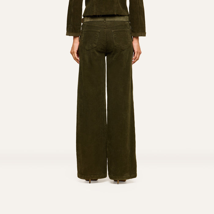 A person stands facing away from the camera against a plain, light background, wearing the Scottie Corduroy Pant—high-rise, dark green wide-leg pants with two back pockets—styled for a look inspired by fall 2026 fashion and paired with a matching jacket.