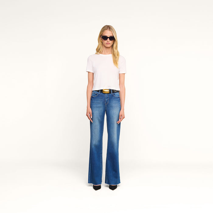 Woman with long blonde hair wearing sunglasses, a white t-shirt, the Scottie Wide-Leg Jean featuring a vintage wash and high-rise flared silhouette, styled with a black belt and black heels, standing against a plain white background.