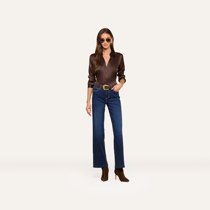 A woman wearing sunglasses, a dark brown long-sleeve shirt, the Scottie Wide-Leg Jean crafted from super stretch denim, a belt, and brown boots stands against a plain light background.