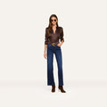 A woman wearing sunglasses, a dark brown long-sleeve shirt, the Scottie Wide-Leg Jean crafted from super stretch denim, a belt, and brown boots stands against a plain light background.