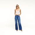 A woman wearing sunglasses, a white tank top, the Scottie Wide-Leg Jean in an indigo wash, and brown shoes stands against a plain white background with her hands in her pockets.