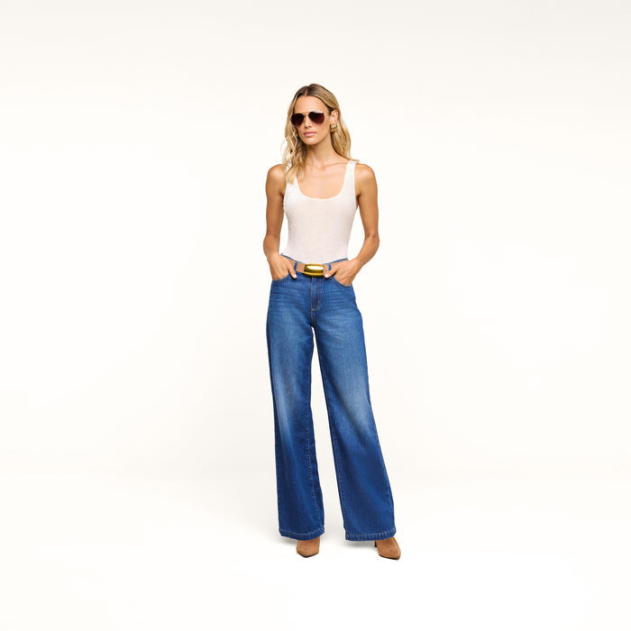 A woman wearing sunglasses, a white tank top, the Scottie Wide-Leg Jean in an indigo wash, and brown shoes stands against a plain white background with her hands in her pockets.